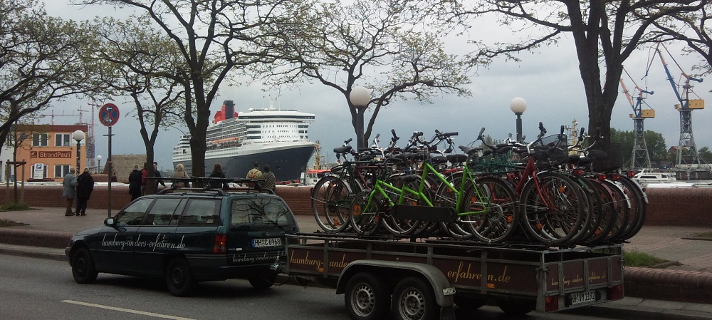 Blick Hafenstraße mit Fahrradtransport, Pkw mit auf dem Anhänger stehenden Fahrrädern. Im Hintergrund die auslaufende Queen Mary 2
